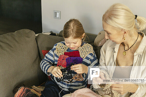 Grandmother looking at granddaughter playing with knitted object sitting on sofa at home