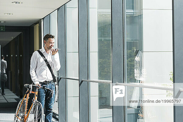 Smiling businessman talking on smart phone and standing with bicycle near window