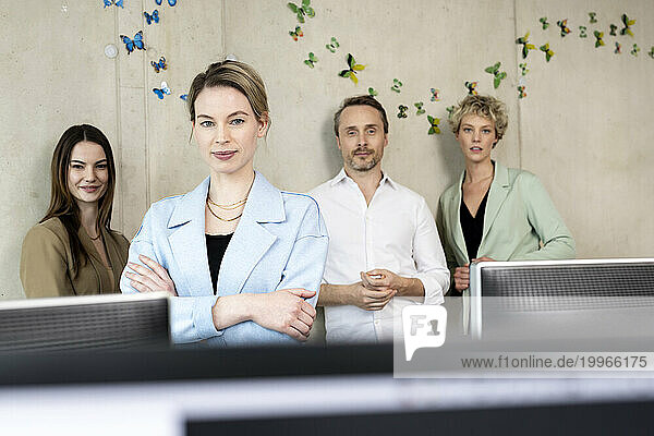 Confident businesswoman with arms crossed by colleagues in workplace