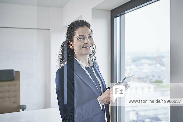 Smiling mature businesswoman standing with tablet PC near glass window