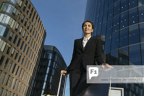 Smiling businesswoman with suitcase standing near buildings in city under sky