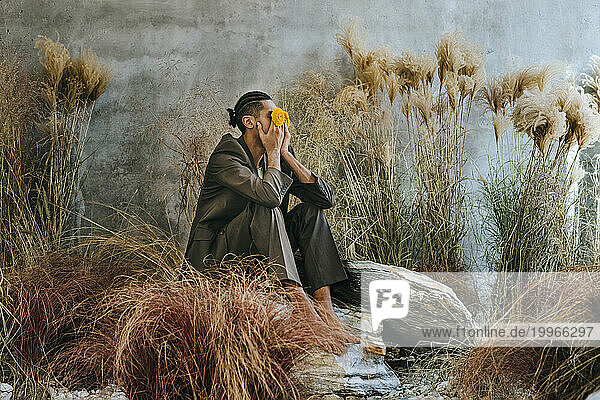 Young businessman covering eyes with flowers and sitting on rock in garden