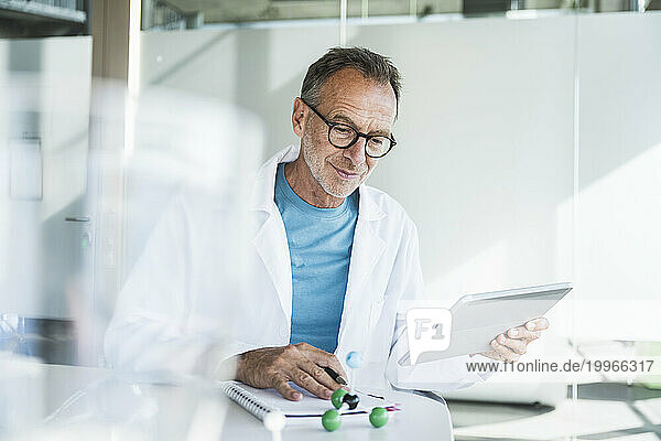 Smiling senior doctor using tablet PC and sitting with note pad at desk