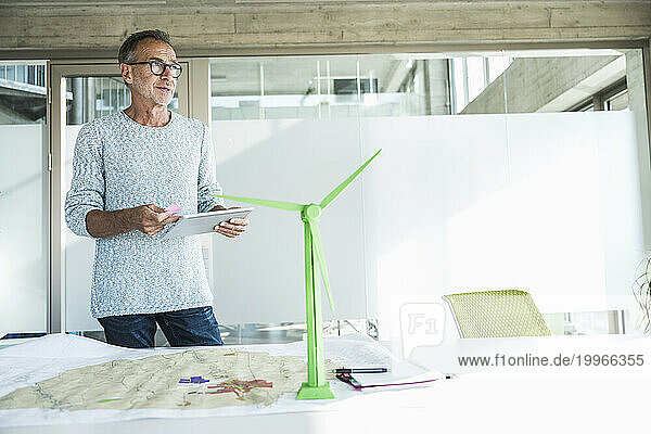 Thoughtful businessman standing with tablet PC near wind turbine model at desk