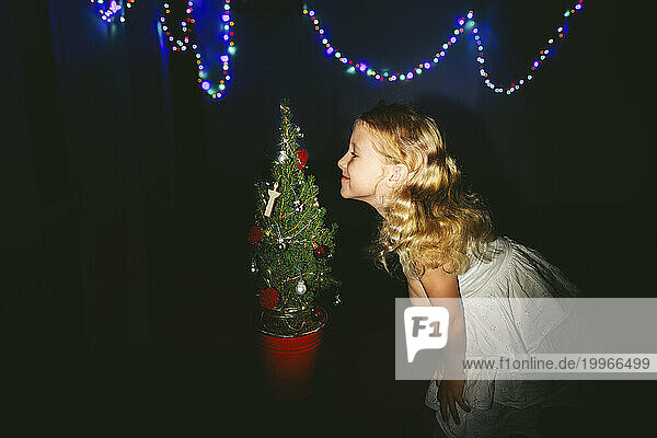 Smiling girl smelling decorated Christmas tree near string lights