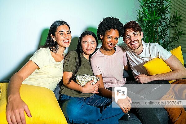 Porträt von multiethnischen Freunden  die auf dem Sofa sitzen und in die Kamera schauen  um einen Film zu sehen