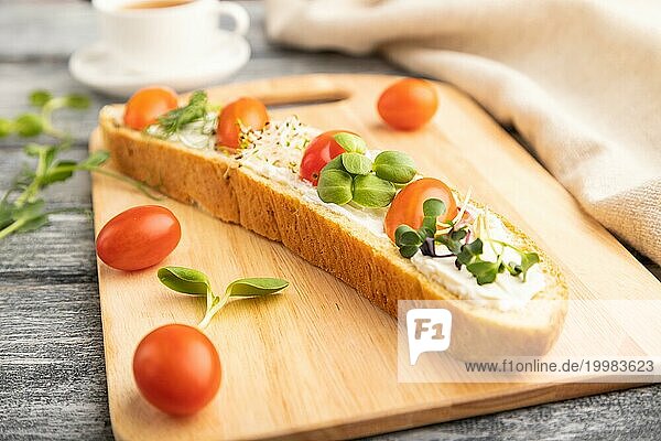 Langes Weißbrot Sandwich mit Frischkäse  Tomaten und Mikrogrün auf grauem Holzhintergrund und Leinenstoff. Seitenansicht  Nahaufnahme  selektiver Fokus