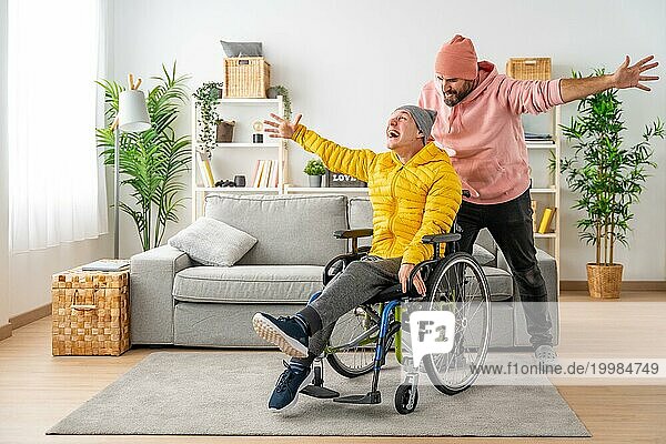 Glücklicher behinderter Mann im Rollstuhl und Freund feiern mit erhobenen Händen zu Hause