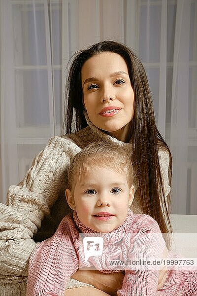 Eine schöne und liebevolle Mutter und eine süße Tochter sitzen in gestrickten Pullovern zusammen im Wohnzimmer