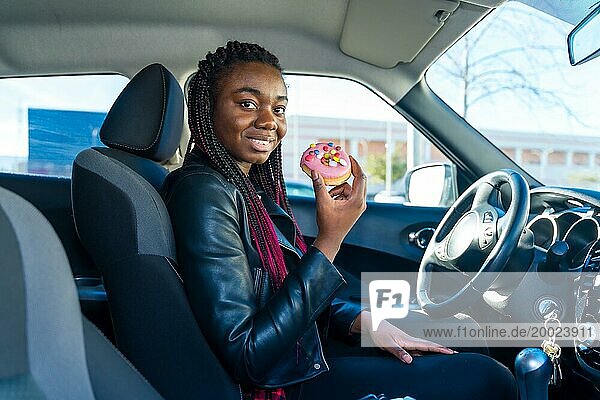 Seitenansicht einer glücklichen afrikanischen Frau  die auf dem Auto sitzend einen Doughnut isst