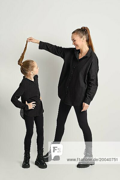 Die schwarz gekleidete Mutter zieht spielerisch am Pferdeschwanz ihrer Tochter  die ebenfalls schwarz gekleidet ist. Das Bild zeigt einen Moment der Freude und Verbundenheit zwischen Mutter und Kind