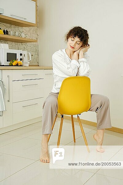 Verträumte junge Frau sitzt mit geschlossenen Augen auf einem Stuhl in der Küche