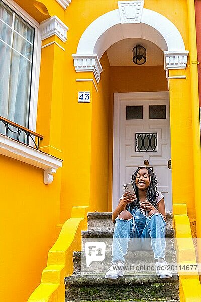 Vertical portrait of a smiling african woman using phone sitting in outside stairs of a yellow colored house
