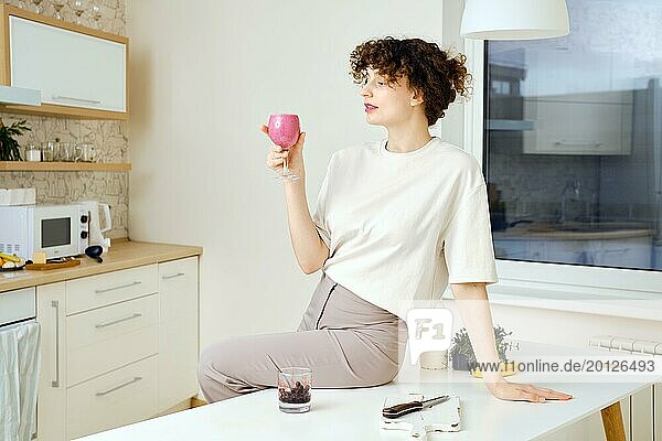 Verträumte und zufriedene junge Frau sitzt auf einem Küchentisch und hält ein Glas Smoothie in der Hand