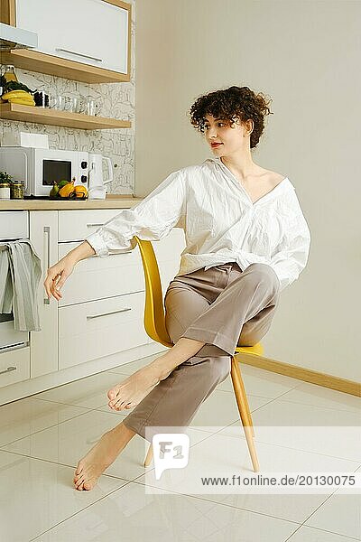 Verträumte junge Frau mit lockigem Haar sitzt in der Küche in eleganter und entspannter Pose