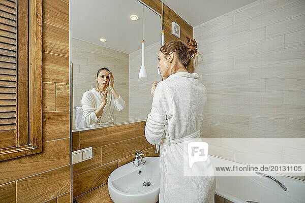 Young woman in white bathrobe washing her face with wash milk standing in front of large mirror in bathroom