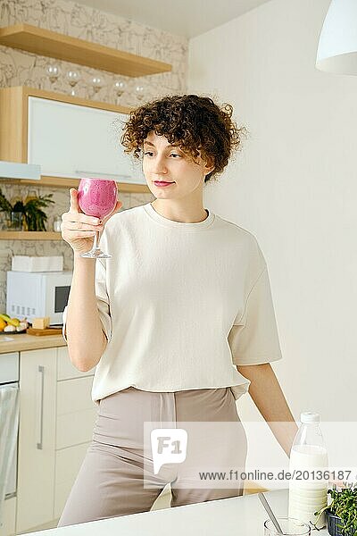 Cheerful woman holds a glass of smoothie in hand which she prepared in her kitchen