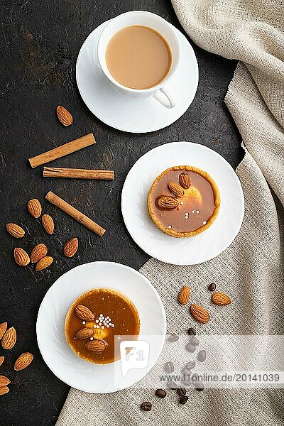 Süße Törtchen mit Mandeln und Karamellcreme mit einer Tasse Kaffee auf schwarzem Betonhintergrund und Leinentextil. Draufsicht  Flat Lay  Nahaufnahme
