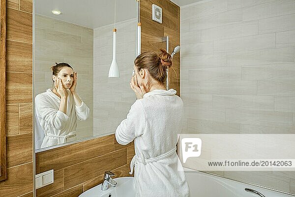 A woman in a white robe is washing her face  standing before a large mirror in a well-lit  contemporary bathroom