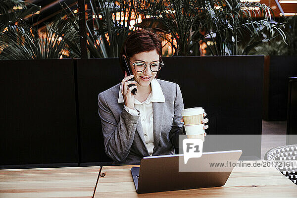 Lächelnde Geschäftsfrau hält Kaffeetasse in der Hand und telefoniert im Café