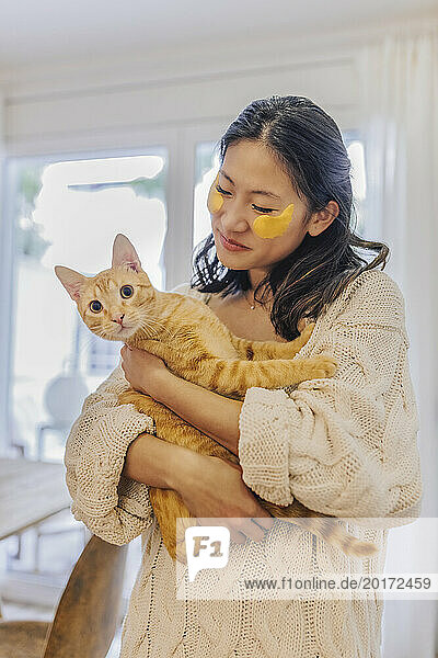 Smiling woman with under eye patches holding cat at home