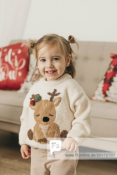 Smiling cute girl wearing Christmas sweater near sofa at home