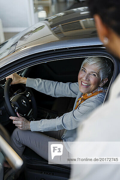 Happy woman sitting in car near salesperson in showroom
