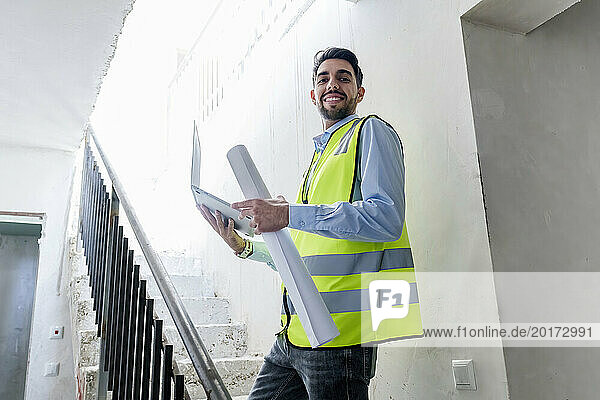 Smiling architect standing with blueprint and laptop at site