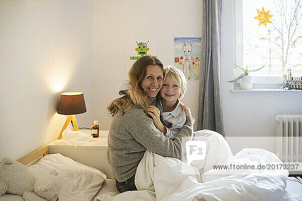 Smiling boy sitting with mother on bed at home