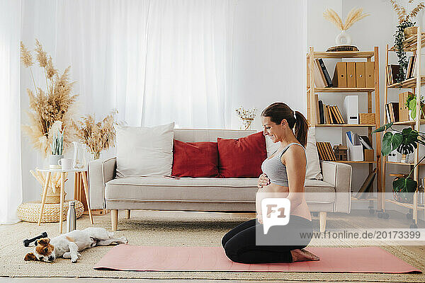 Smiling pregnant woman kneeling with hands on stomach looking at dog in living room