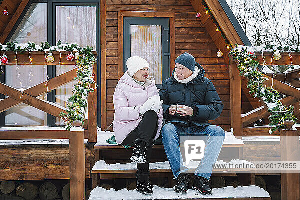 Mature couple sitting and having tea on porch near log cabin