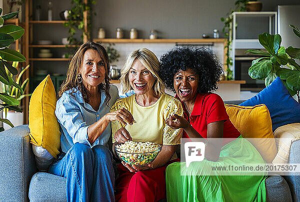 Cheerful friends sitting on sofa and sharing popcorn at home