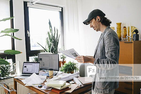 Side view of male architect analyzing blueprint while standing by desk in home office