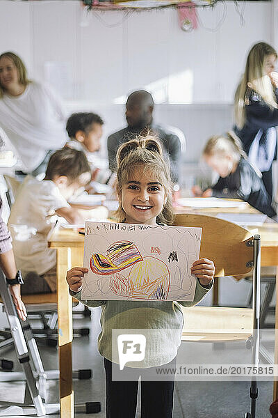 Portrait of smiling girl showing drawing while standing near bench in classroom at kindergarten