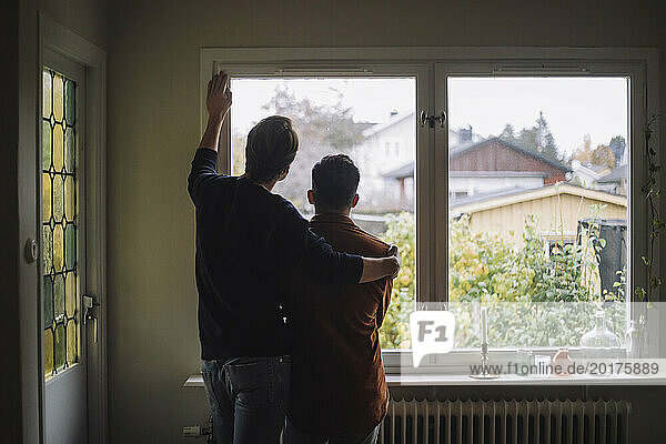Rückansicht eines Mannes mit einem Arm um ein schwules Paar, das zu Hause durch das Fenster schaut