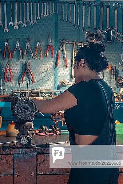 Mechanikerin  die eine Lichtmaschine an einer Werkbank in einer Autowerkstatt repariert  trägt einen schwarzen Overall und ein schwarzes Hemd  eine komplette Werkzeugtafel im Hintergrund mit Bokeh Effekt  traditionelle Männerberufe von Mixed race latino woman
