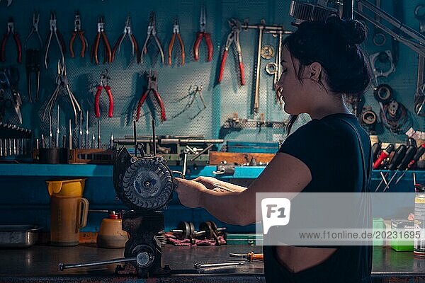 Gemischtrassige hispanische junge Mechanikerin  trägt schwarzen Overall  konzentriert sich auf ein Motorradteil in einer Werkstatt mit einem Werkzeugschrank  eine komplette Werkzeugtafel im Hintergrund mit Bokeh Effekt  traditionelle Männerberufe von gemischtrassiger Latino Frau