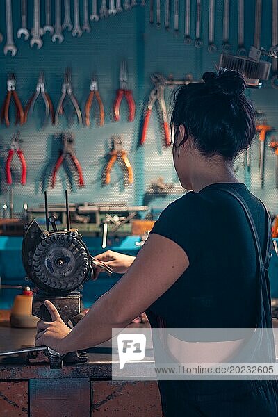 Weibliche hispanische Mechanikerin arbeitet an einer Werkbank in einer gut ausgestatteten Werkstatt von hinten  eine komplette Werkzeugtafel im Hintergrund mit Bokeh Effekt  traditionelle männliche Berufe von Mixed race latino woman
