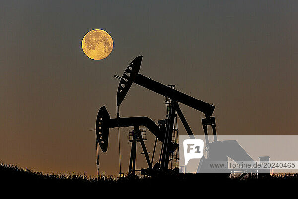 Silhouette of two pumpjacks with a glowing warm sky and full moon  West of Airdrie; Alberta  Canada