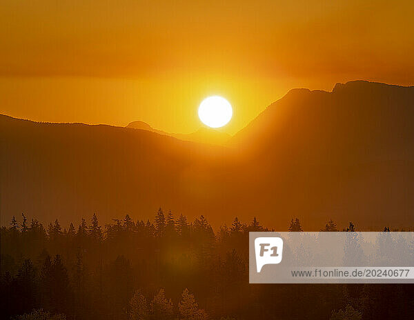 A bright sun with orange sky and golden sunlight glowing at twilight over the silhouetted mountain peaks and forest; British Columbia  Canada