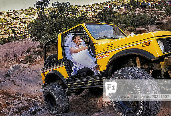Bride hangs on for a four-wheel drive wild ride over boulders and rough slick rock trails near Moab  Utah. The Bureau of Land Managements designates specific trails for off-road vehicle riders like this who although dressed in traditional white  wants to be married in an untraditional way in the wilderness during a Jeep Safari. Most riders stick to BLM's loosely enforced straight-and-narrow rules are plentiful  but thousands more disregard the rules  answering the call of their combustion engines to chart new paths through roadless areas which had great ecological consequences; Utah  United States of America