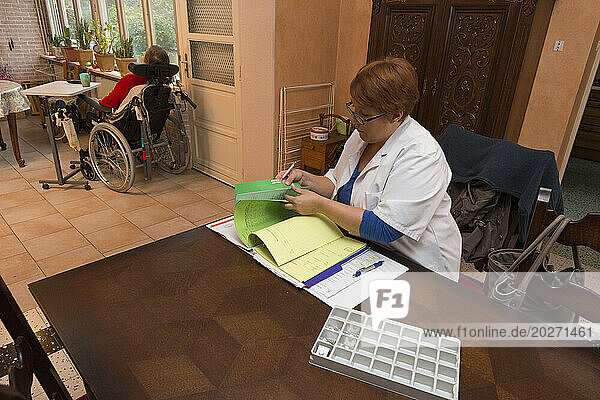HAD (Hospitalization At Home). Follow-up of an elderly patient at her home by a caregiver. Nursing assistant filling out the transmission book with the elderly and disabled patient installed in her wheelchair in the background.