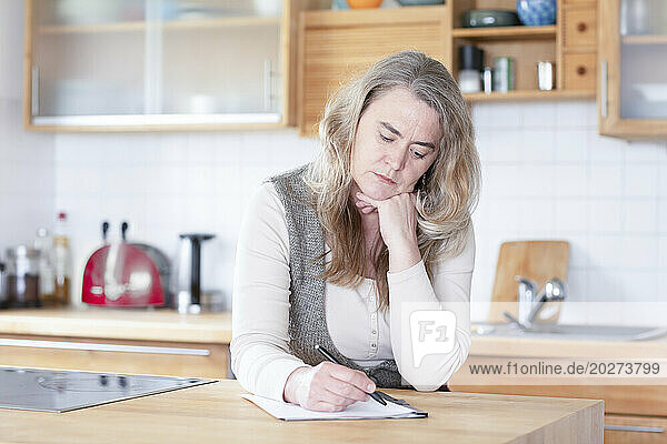 Mature woman writing on paper in kitchen at home