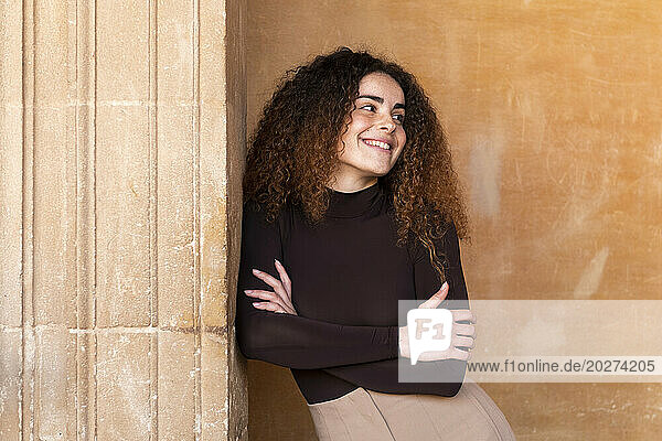 Smiling young woman with arms crossed leaning on wall