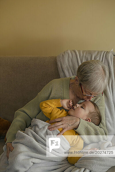 Grandmother kissing grandson sitting on sofa at home