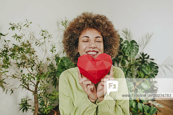 Happy woman holding red paper heart near plants
