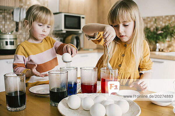 Sisters dipping Easter eggs in colored dye at home