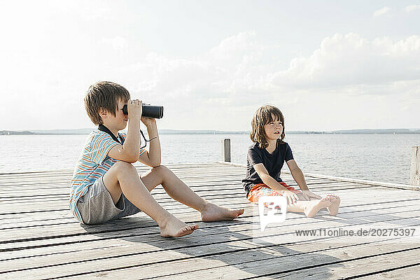 Boy looking through binoculars sitting with brother on pier at lake