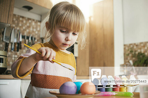 Cute girl coloring Easter eggs with paintbrush at home