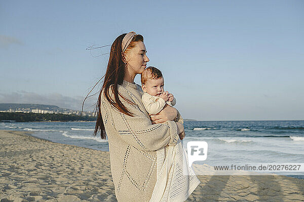 Thoughtful woman holding daughter and standing near sea at beach